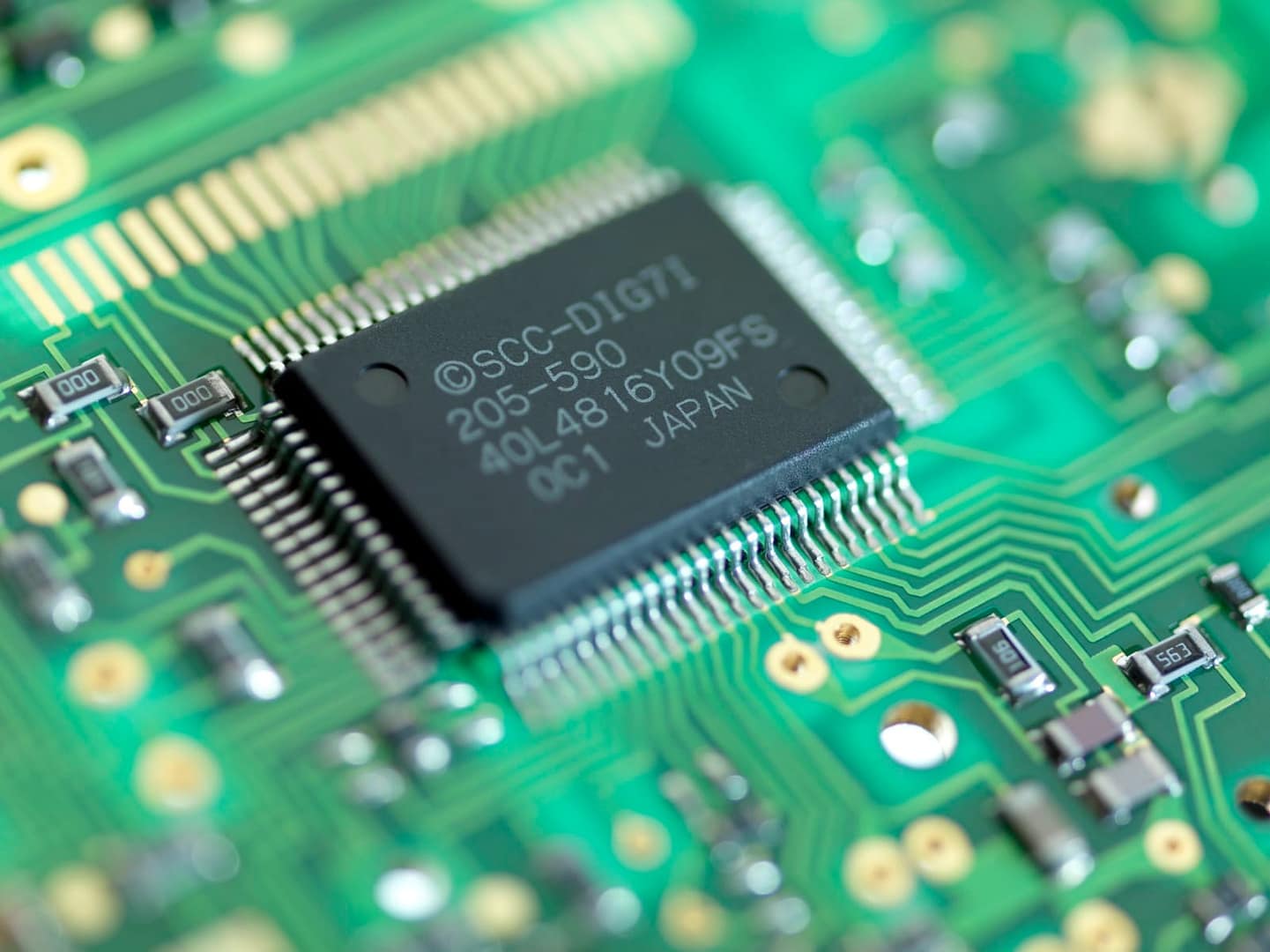 Close-up of a green circuit board featuring a black integrated circuit chip with pins extending outwards. The board has intricate gold pathways.