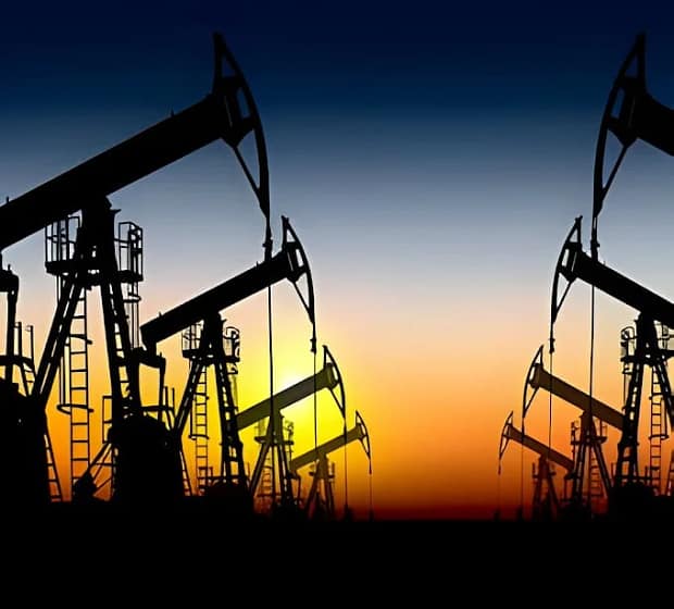 Silhouetted oil pumps line up against a vivid sunset, blending orange, yellow, and blue hues. The scene is industrial yet serene, evoking energy production.